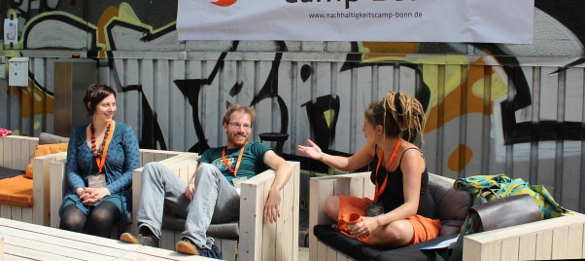 Drei Teilnehmende des NachhaltigkeitsCamps sitzen im Freien auf Holz-Sesseln und unterhalten sich.