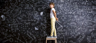 Ein Mädchen steht auf einem Hocker vor einer bildfüllenden Tafel mit Formeln und Notizen angeschrieben
