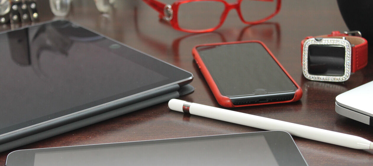 Tablets, Smartphone und Smartwatch liegen mit einer Brille und verschiedenen Büroutensilien auf einem Schreibtisch. 