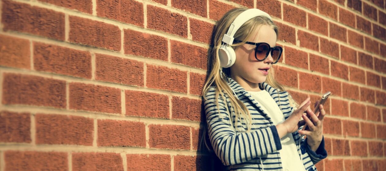 Ein junges Mädchen lehnt mit Kopfhörern auf und einem Handy in der Hand an einer Backsteinwand