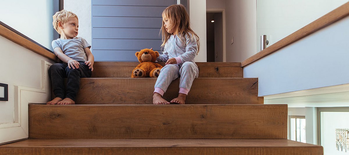 Zwei Kinder im Kindergartenalter sitzen weit auseinander auf einer Treppe und haben einen Teddybär in ihrer Mitte.