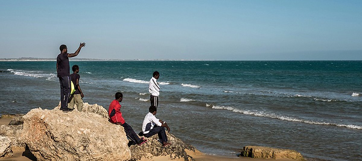 Fünf afrikanische Männer stehen und sitzen auf Felsen am Strand und schauen auf das Meer.