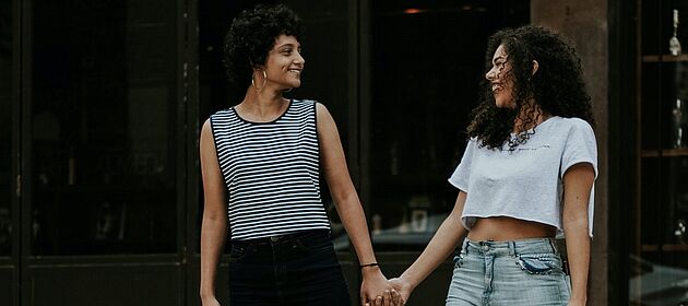 Zwei queere Personen halten sich an der Hand und lachen