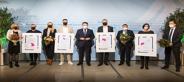 Auf dem Foto sind die Gewiner/-innen des  Wettbewerbs "Innovatives Netzwerk 2021" zu sehen. Sie stehen vor einer Wand und halten ihre Auszeichnung sowie Blumensträuße in der Hand. 