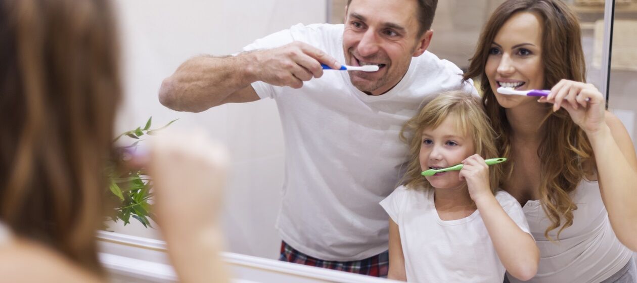 Ein Mädchen steht mit ihren Eltern vor dem Badezimmerspiegel. Alle drei putzen ihre Zähne.