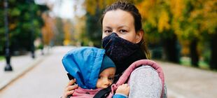Frau mit Mundschutz trägt ein Baby mit Kapuze auf dem Arm