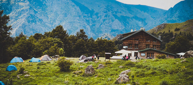 Jugendliche zelten auf einer Wiese vor einem Landhaus mit Alpenpanorama