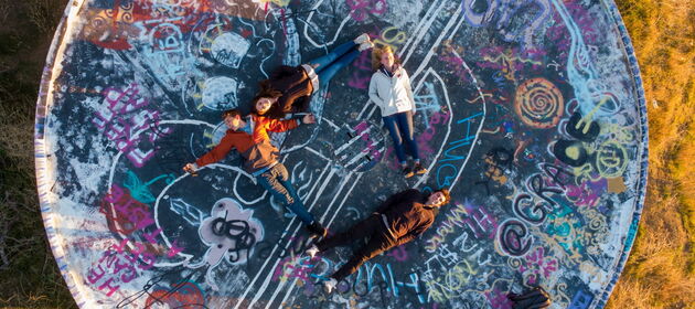 Vier Jugendliche liegen unter freiem Himmel auf dem Rücken auf einer mit Graffitti versehenen Fläche