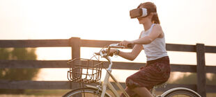 Ein Mädchen sitzt mit einer VR-Brille auf einem Fahrrad und fährt an einem Holzzaun vorbei.  