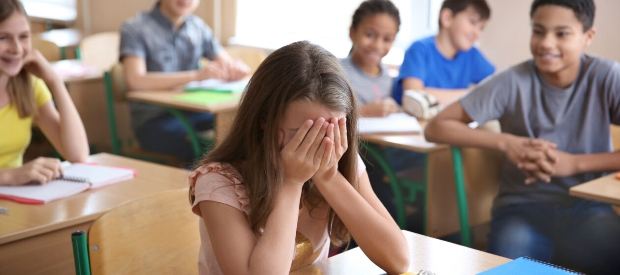 Eine Schülerin verbirgt im Klassenraum beschämt ihr Gesicht hinter ihren Händen, ihre Mitschüler lachen sie aus