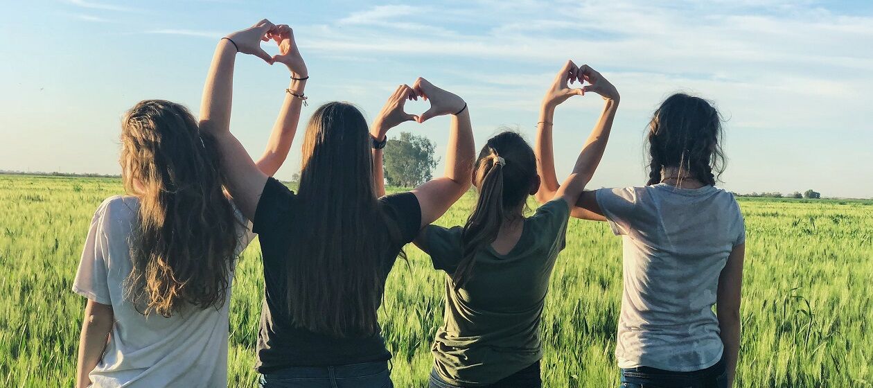 Vier Mädchen stehen in einer Reihe vor einem Kornfeld und bilden mit ihren Händen Herzen