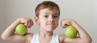 Ein Junge schaut konzentriert und hat zwischen seinen Ober- und Unterarmen zwei grüne Äpfel geklemmt