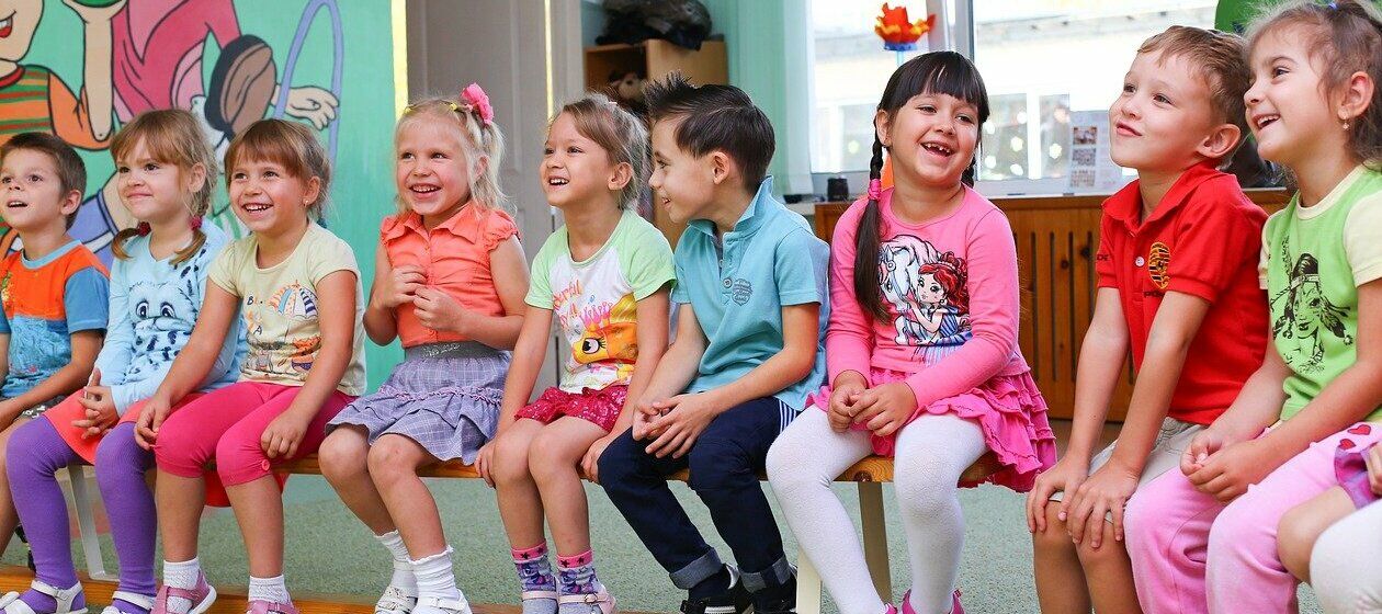 Eine Gruppe Kinder sitzt auf einer Bank im Kindergarten. Alle lachen.