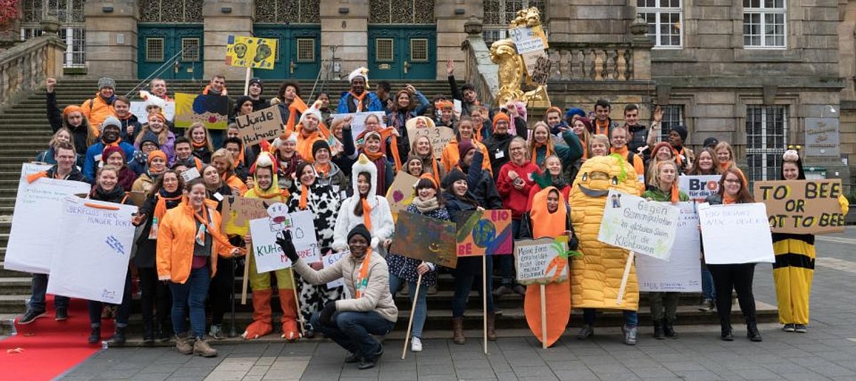 Teilnehmende von Brot für die Welt Jugend stehen mit Schildern und Kostümen in einer Gruppe, bei einer Straßenaktion für gerechte Landwirtschaft.
