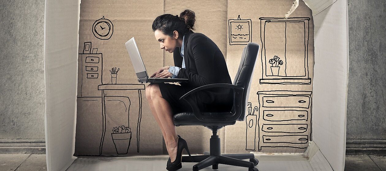Eine Frau sitzt zusammengekauert auf einem Bürostuhl und arbeitet am Laptop, dabei sitzt sie in einem seitlich aufgestellten Karton, in den die Einrichtung eines Hauses gemalt ist