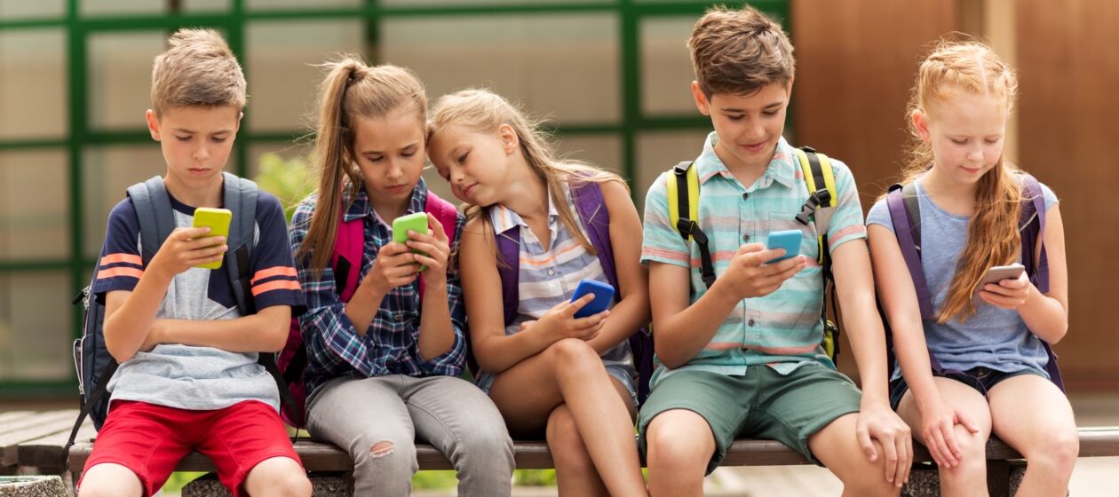 Fünf Schüler und Schülerinnen mit Rücksäcken sitzen auf einer Bank und schauen alle auf ein Handy