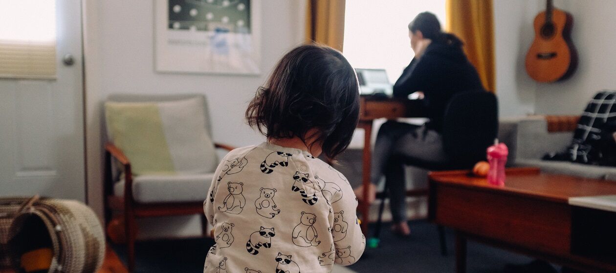 Kleinkind von hinten mit Frau, die im Hintergrund am Schreibtisch vor einem PC sitzt
