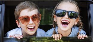 Zwei Kinder mit Sonnenbrille schauen aus dem heruntergelassenen Fenster eines Autos