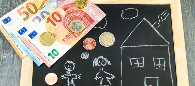 Auf einer Schiefertafel mit einer Zeichnung von einem Haus und Kindern liegt Geld. Das Bild symbolisiert Baukindergeld.  