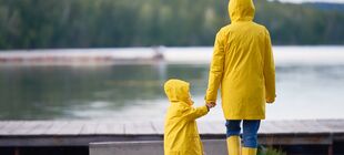 Eine erwachsene Person und ein Kleinkind stehen an einem See und halten sich an der Hand, beide tragen gelbe Regenmäntel