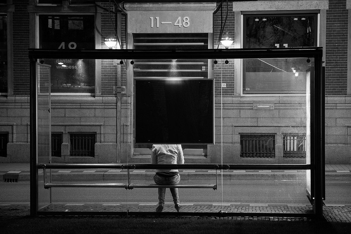 Der Rücken einer Frau, die alleine an einer Bushaltestelle in einer dunklen Straße sitzt.