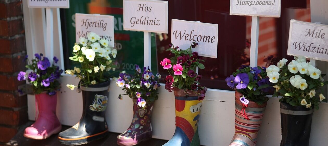 Mit Blumen bestückte Kindergummistiefel stehen vor einem Fenster. In den Stiefel stecken weiße Schilder mit Willkommensgrüßen in verschiedenen Sprachen.