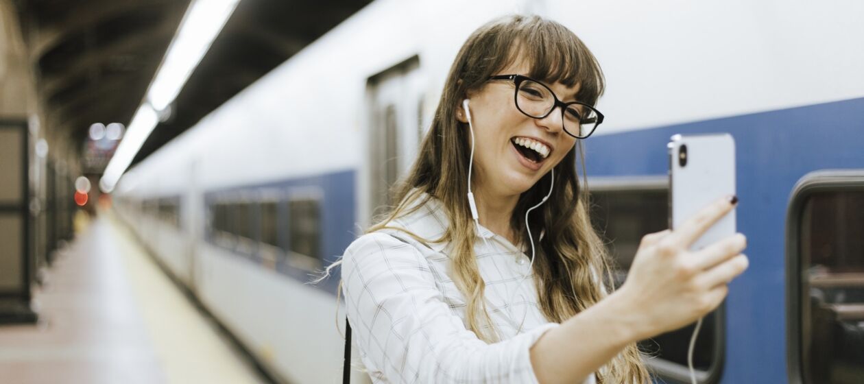 Junge Frau an Bahnhof hat Kopfhörer auf und lacht in ihr Smartphone