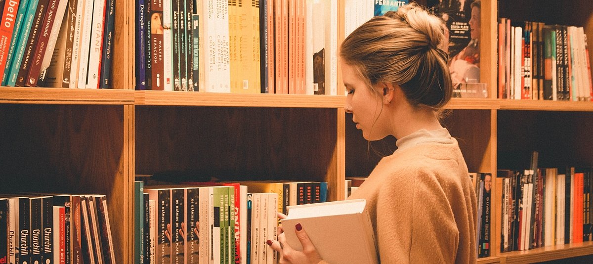 Eine Jugendliche steht mit einem Buch in der Hand in einer Bibliothek vor einem Bücherregal