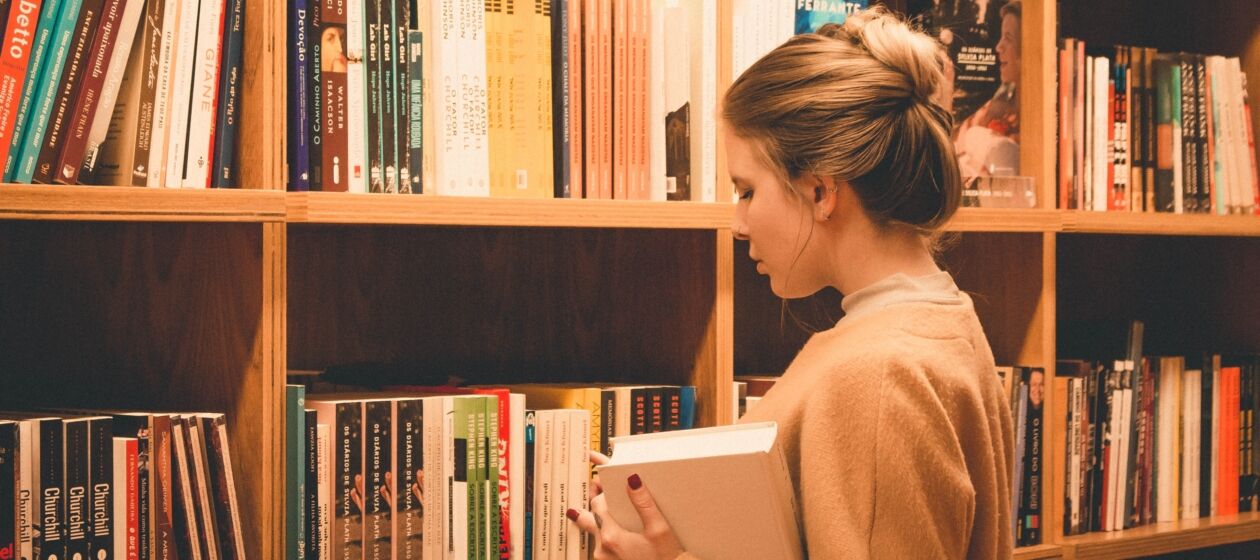 Eine Jugendliche steht mit einem Buch in der Hand in einer Bibliothek vor einem Bücherregal