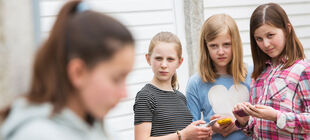 Drei Mädchen halten ihre Handys in der Hand und schauen verächtlich auf ein anderes Mädchen, das verschwommen im Vordergrund zu sehen ist