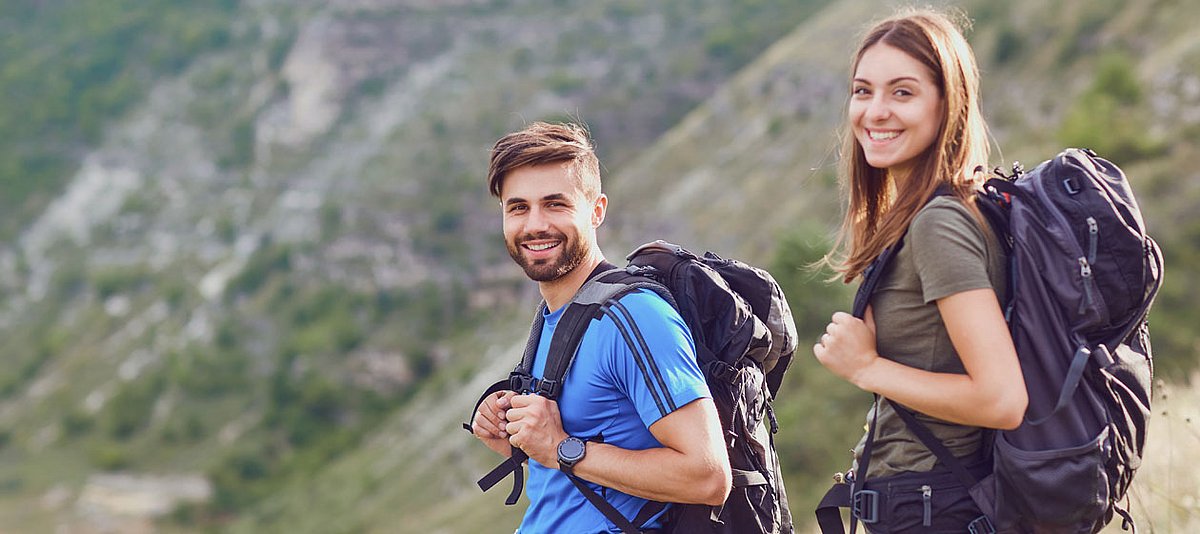 Ein junges Paar mit Rucksäcken läuft durch ein Gebirge und lächelt glücklich