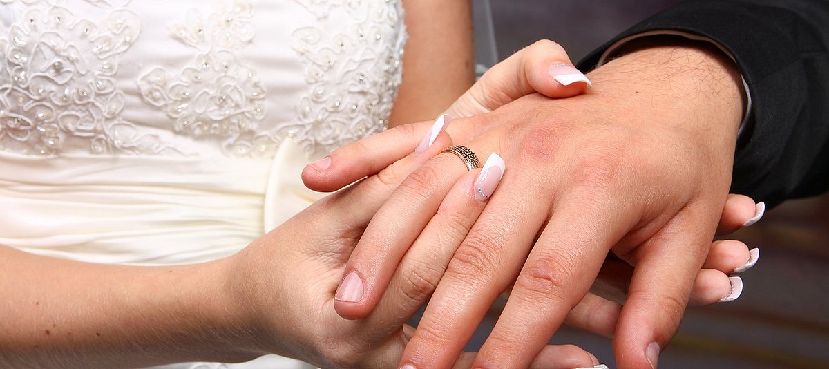 Die Braut steckt ihrem Bräutigam den Ehering an den Finger und hält seine Hand.