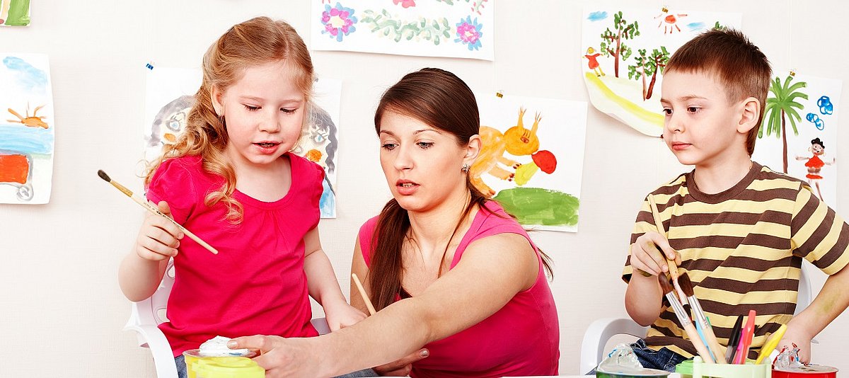eine Betreuerin sitzt in der Mitte zweier Kleinkinder, mit denen sie malt