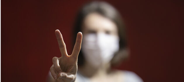 Frau mit Mundschutz zeigt ein Victory Zeichen mit ihrer Hand