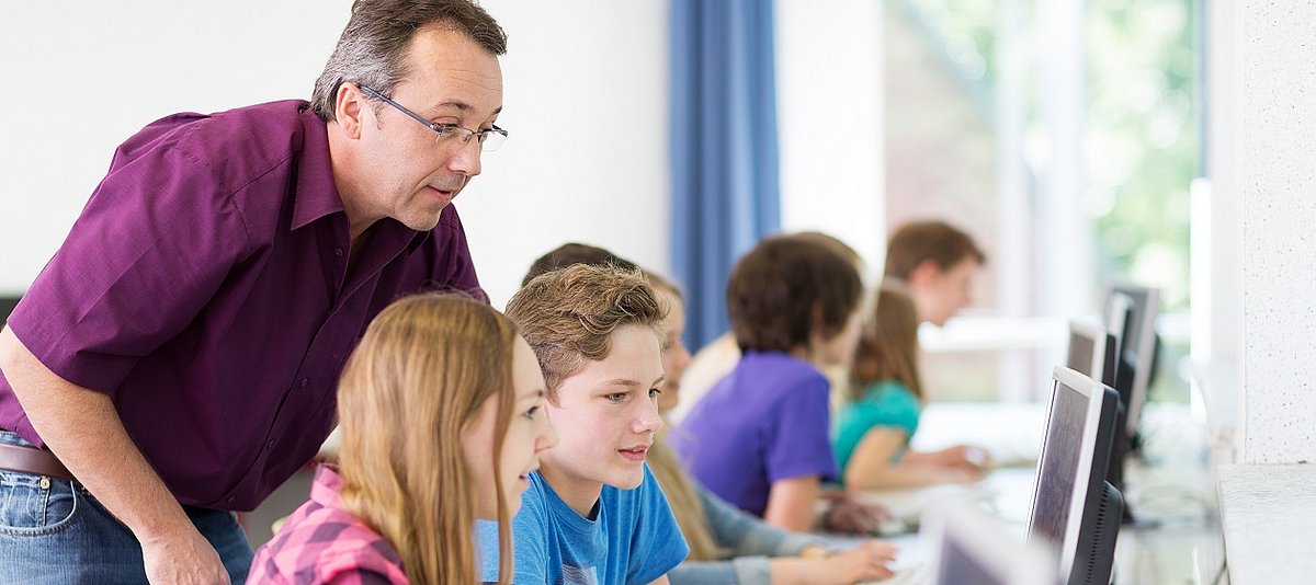 Im Klassenzimmer sitzen Schüler an den Computern. Ein Lehrer steht hinter ihnen und erklärt etwas.