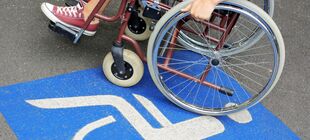 Eine jugne Frau im Rollstuhl steht auf einem Behinderten-Parkplatz, der durch ein blau-weißes Schild auf dem Asphalt gekennzeichnet ist