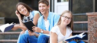 Drei Jugendliche sitzen vor der Schule auf einer Treppe, halten Hefte in der Hand und lachen