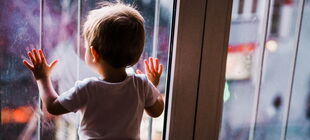 Ein kleines Kind steht in einem Raum an einer Fensterfront, die vergittert ist, und schaut nach draußen