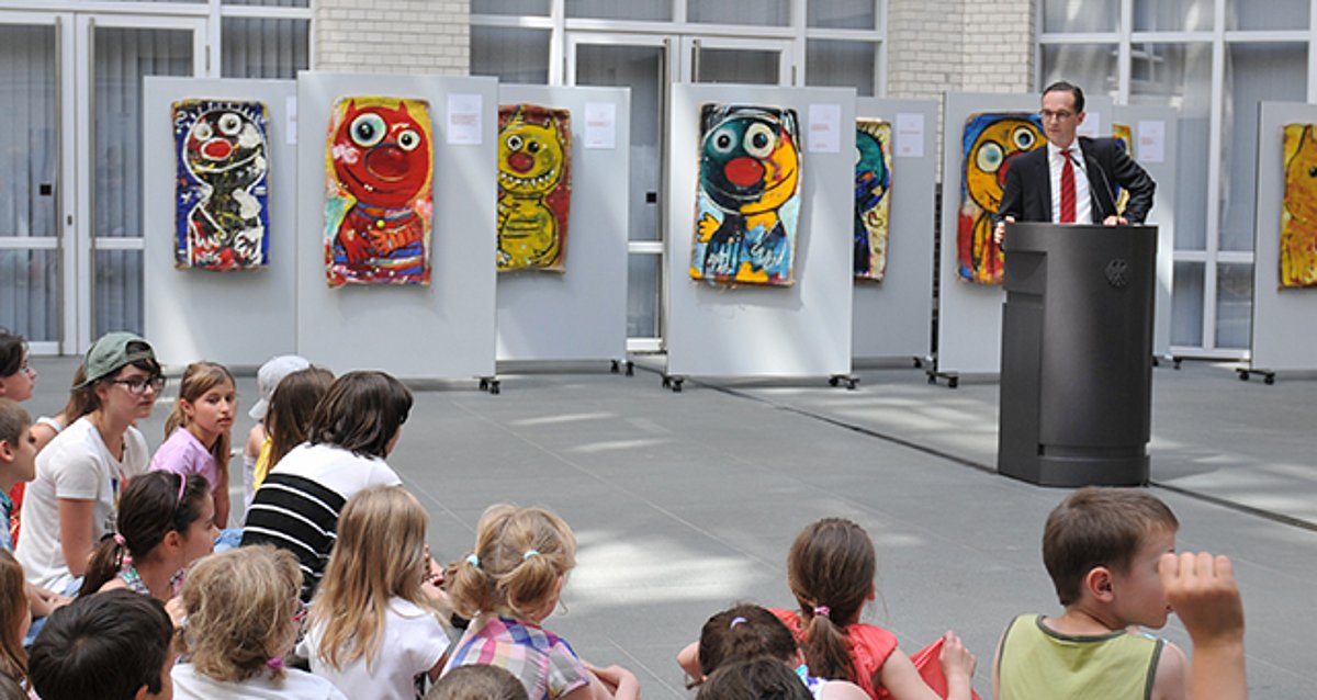 Bundesminister Maas steht am Rednerpult vor den ausgestellten Bildern der kleinen Monster und spricht zu Schulkindern