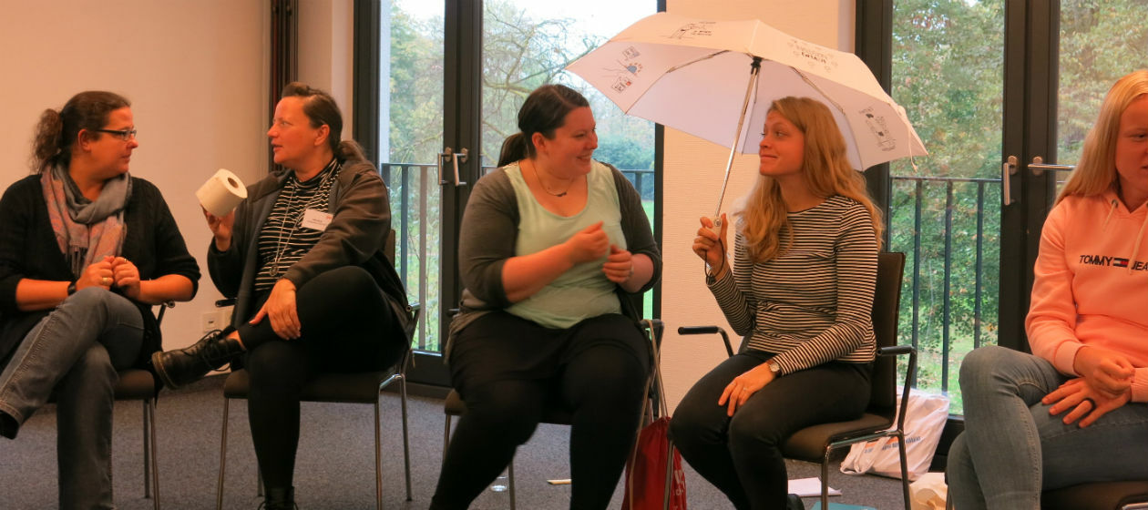 Vier Frauen sitzen nebeneinander und unterhalten sich. Eine hält einen Regenschirm, eine andere eine Toilettenpapierrolle.