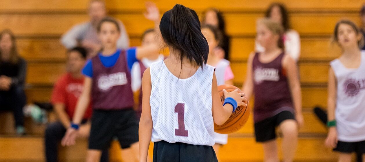 Mädchen spielen Basketball in einer Halle, im Hintergrund sitzen wenige Zuschauer auf einer Tribüne