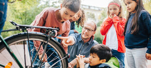 Ein Mann hockt mit einer Gruppe Kinder um ein Fahrrad und erklärt das Rücklicht.