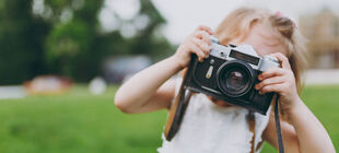 Ein kleines Mädchen auf einer Wiese hält aich eine große Kamera vor ihr Gesicht