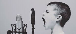 Ein Junge mit sehr kurzem Haar spricht mit aufgerissenem Mund in ein Studiomikrofon.