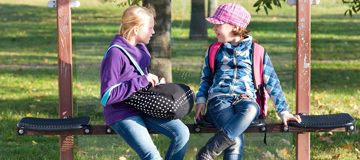 Zwei Mädchen sitzen an einer Bushaltestelle mit Rucksack und Schultasche..