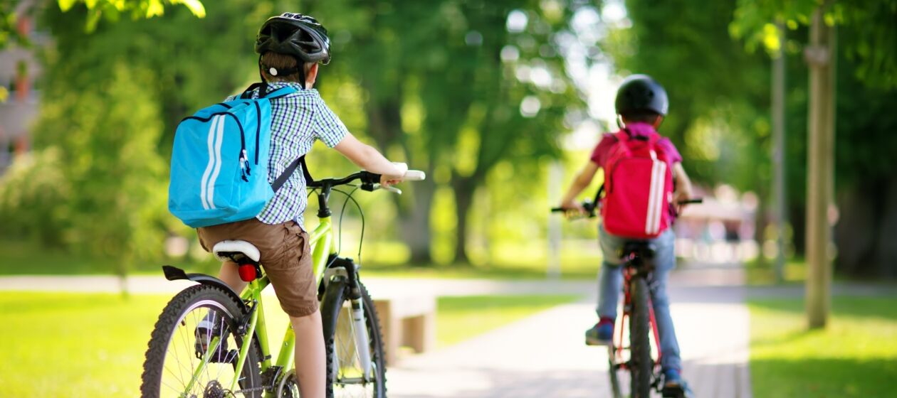 Zwei Schüler mit Rücksäcken und Fahrradhelmen fahren auf einem Fahrradweg, gesäumt von einer Wiese und Bäumen