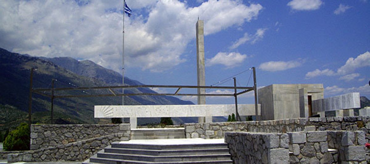 Eine in Naturstein eingefasste Terrasse mit Stele und griechischer Fahne vor einer Bergkulisse, 