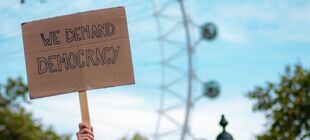Eine Hand hält ein Pappschild zum Himmel, das die Aufschrift „We demand democracy“ trägt. Im Hintergrund ein Riesenrad.