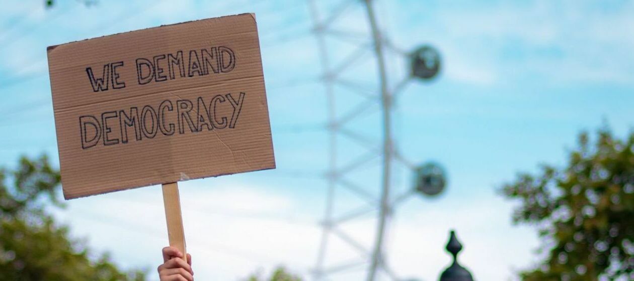Eine Hand hält ein Pappschild zum Himmel, das die Aufschrift „We demand democracy“ trägt. Im Hintergrund ein Riesenrad.