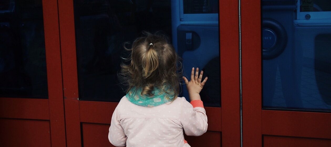 Ein Kind von hinten fotografiert lehnt an eine Tür mit einem Fenster, eine Hand am Fenster